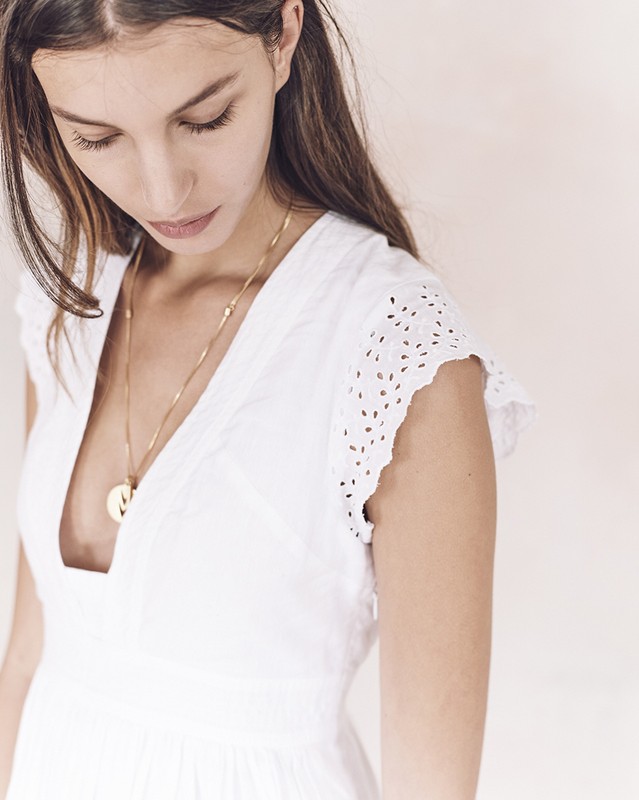 Young woman gazes downward while posing in a white dress with a gold necklace. The image is shot in a photo studio setting.