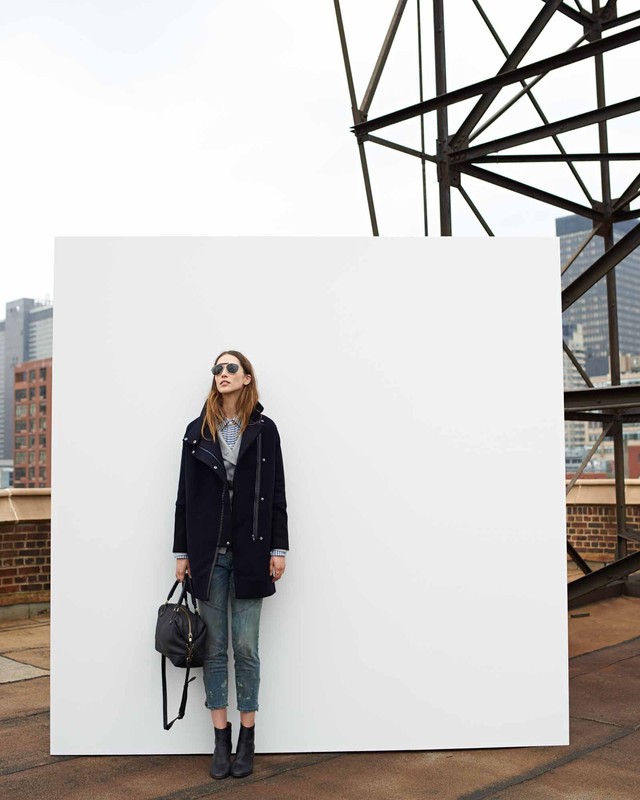 Model wearing dark coat, denim jeans, and sunglasses standing outdoors holding a black handbag against a white wall.