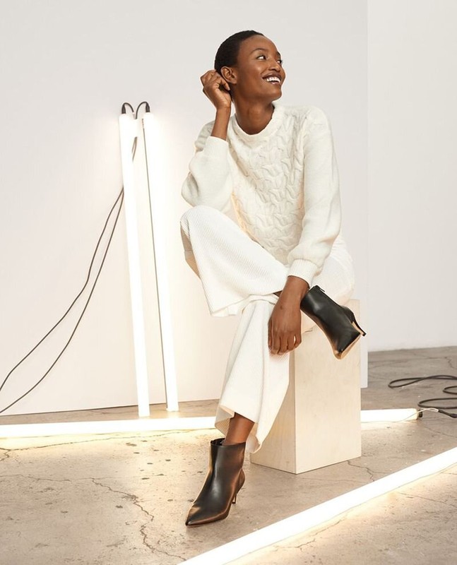 A Black female fashion model smiles while sitting on a white cube in a cable knit sweater, wide leg pants and black stiletto heel booties.
