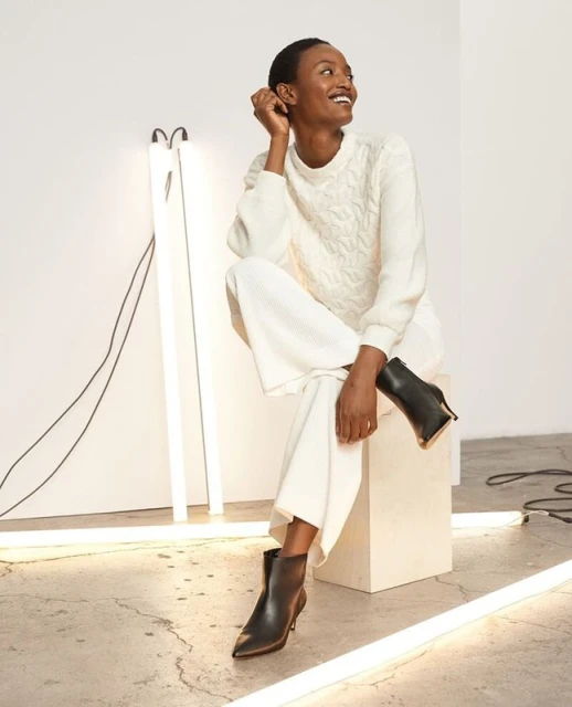 A Black female fashion model smiles while sitting on a white cube in a cable knit sweater, wide leg pants and black stiletto heel booties.