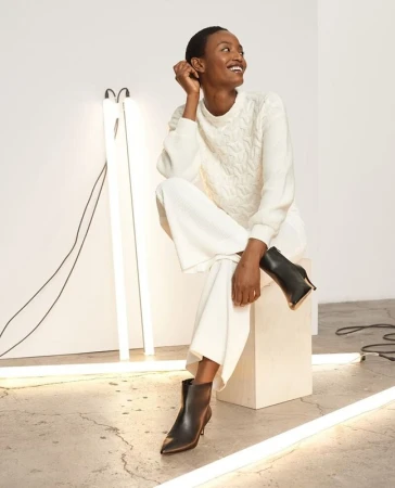 A Black female fashion model smiles while sitting on a white cube in a cable knit sweater, wide leg pants and black stiletto heel booties.
