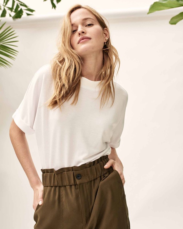 Blonde woman in a white t-shirt and olive pants poses in front of a white wall with green tropical plants. Fashion portrait.