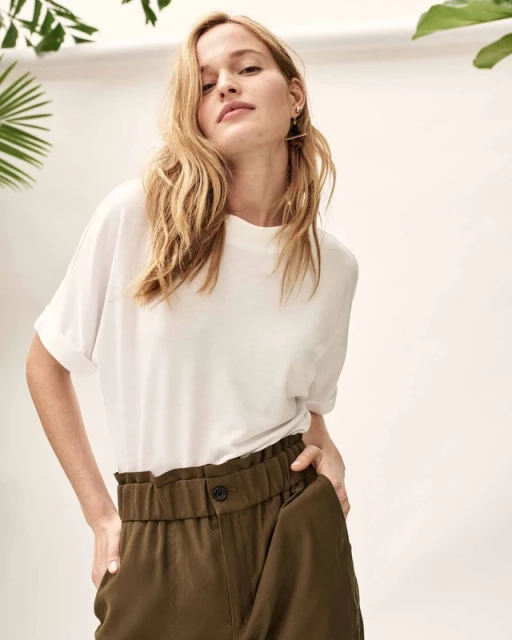 Blonde woman in a white t-shirt and olive pants poses in front of a white wall with green tropical plants. Fashion portrait.