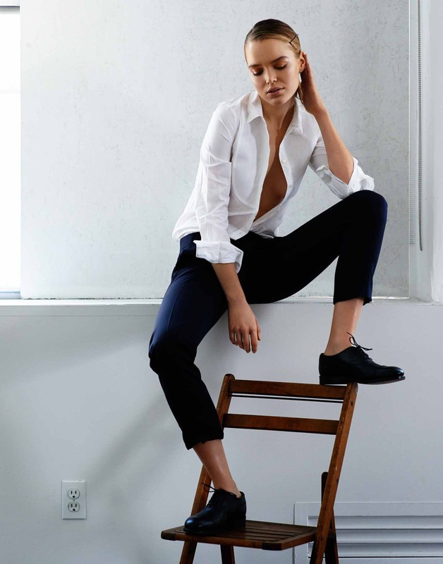 A stylish young woman with blonde hair poses on a chair in a white shirt and dark pants, set against a minimalist white background indoors.