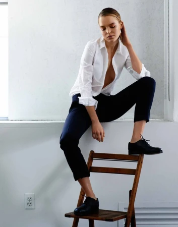 A stylish young woman with blonde hair poses on a chair in a white shirt and dark pants, set against a minimalist white background indoors.