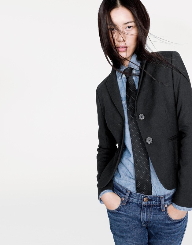 Portrait of an Asian woman in a black blazer, denim shirt, and dark tie against a plain white studio background.