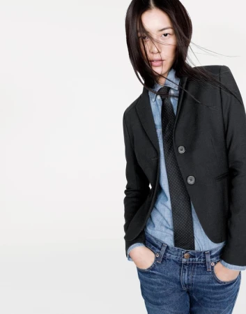 Portrait of an Asian woman in a black blazer, denim shirt, and dark tie against a plain white studio background.