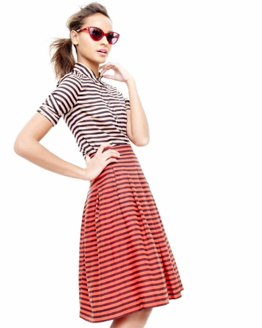 A fashionable adult female poses against a white backdrop, wearing red sunglasses, a striped top, and a red striped skirt.