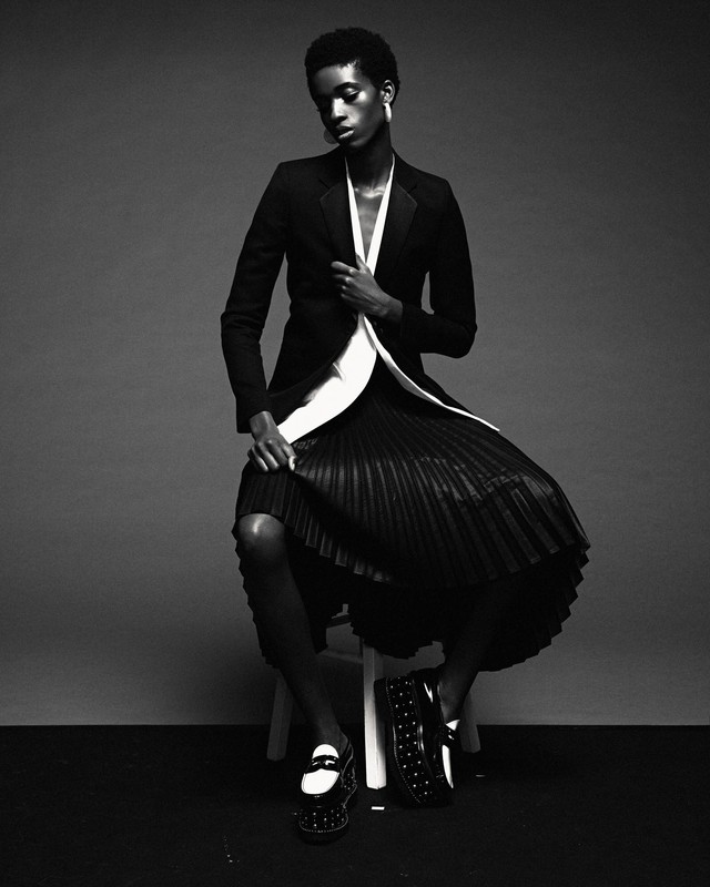 A black and white fashion portrait of a woman with short hair in a blazer and pleated skirt sitting on a white stool.