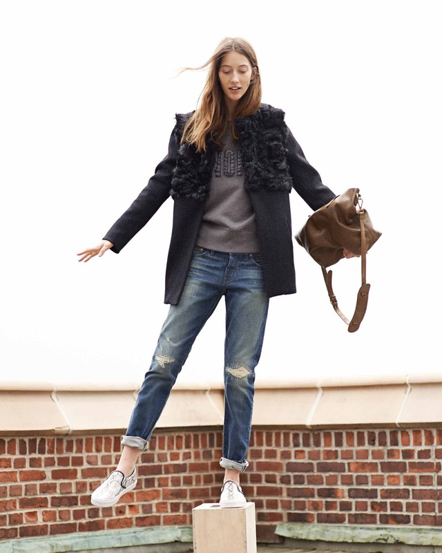 Full-length photo of a female model standing on a block against an outdoor brick wall holding a brown leather handbag.