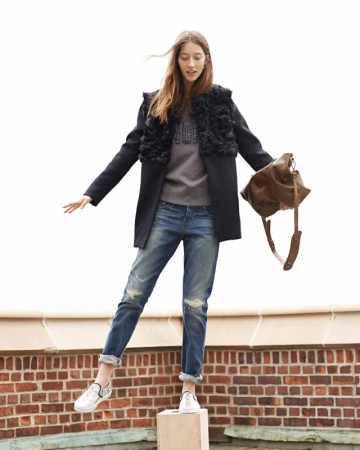 Full-length photo of a female model standing on a block against an outdoor brick wall holding a brown leather handbag.