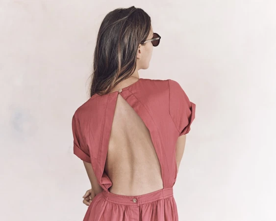 Woman wearing a dusty rose open back dress and sunglasses photographed from behind against a white wall.