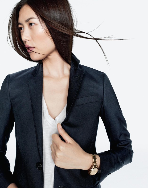 Portrait of an adult woman with long dark hair wearing a navy blue blazer and gold watch, posing in a white studio setting.