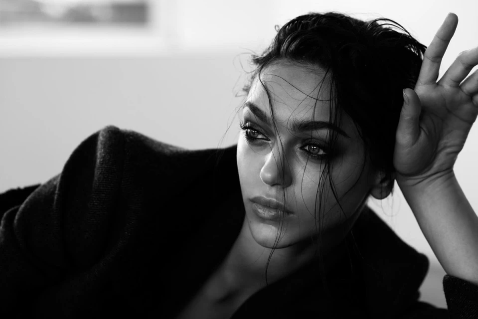 Black and white portrait of a woman with dark hair and eyes, wearing a dark coat, with a soft light creating dramatic shadows.