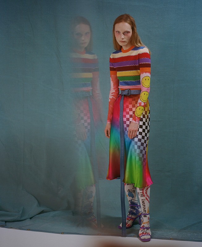 A slender woman with auburn hair poses in a colorful striped top, checkerboard and rainbow skirt, patterned tights, and platform heels against blue.