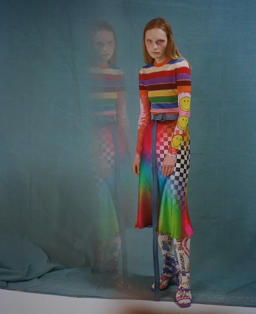 A slender woman with auburn hair poses in a colorful striped top, checkerboard and rainbow skirt, patterned tights, and platform heels against blue.