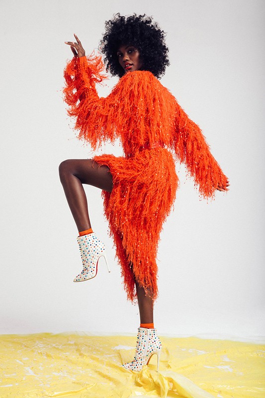 Black woman with afro wearing a fringed orange dress and white studded high heels against a white background.