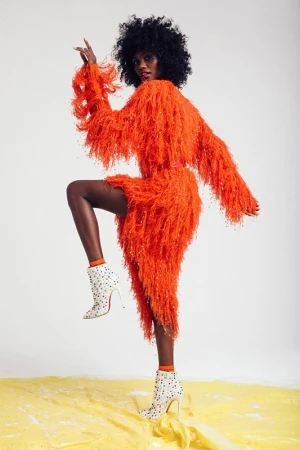 Black woman with afro wearing a fringed orange dress and white studded high heels against a white background.