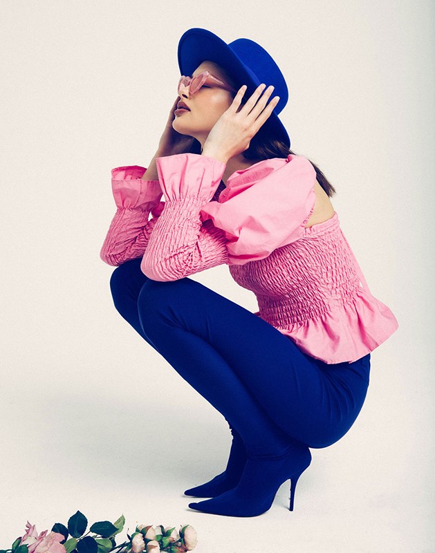 Adult female fashion model in sunglasses and blue hat poses in pink top and blue pants and boots in a photo studio setting with roses.