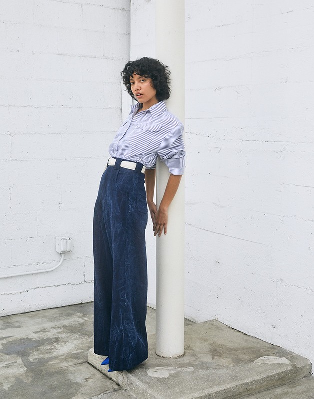 A young woman with curly hair poses in a studio, wearing a checkered shirt and wide-leg denim pants with bright blue platform shoes.