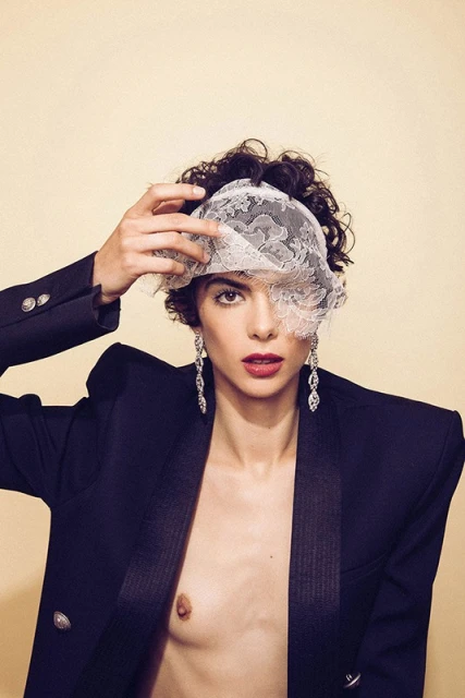 Glamorous woman with dark hair and a lace veil poses in a black blazer, with dangling earrings and red lipstick against a beige backdrop.