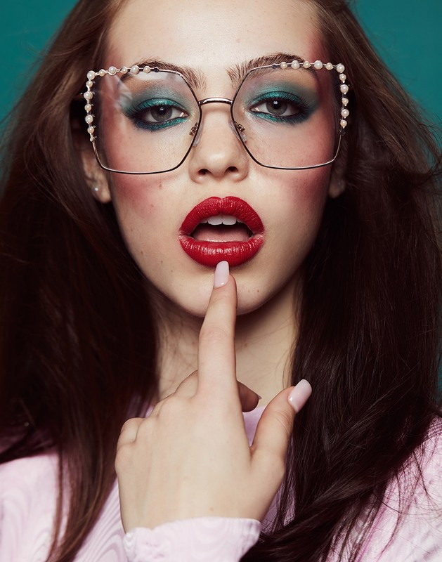 Eye-level portrait of a woman with dark hair, pearl glasses, and red lipstick, posing against a solid teal background in a beauty shot.