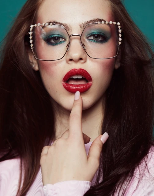 Eye-level portrait of a woman with dark hair, pearl glasses, and red lipstick, posing against a solid teal background in a beauty shot.