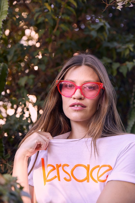 A woman with long brown hair wearing pink sunglasses and a Versace t-shirt poses in front of green foliage outdoors.