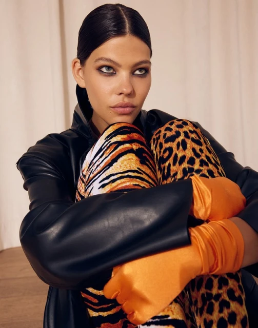 Adult female model with slicked-back hair wears a black leather jacket, bright orange gloves, and animal print pants in a studio setting.