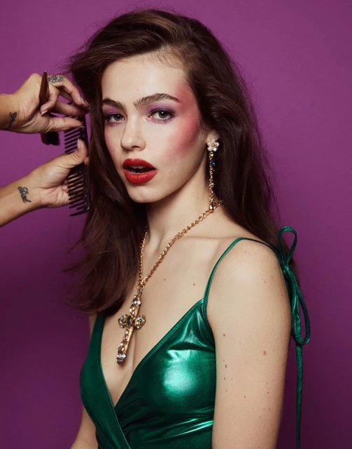 A young woman with dark hair poses with elaborate makeup and jewelry while a stylist combs her hair against a purple background.