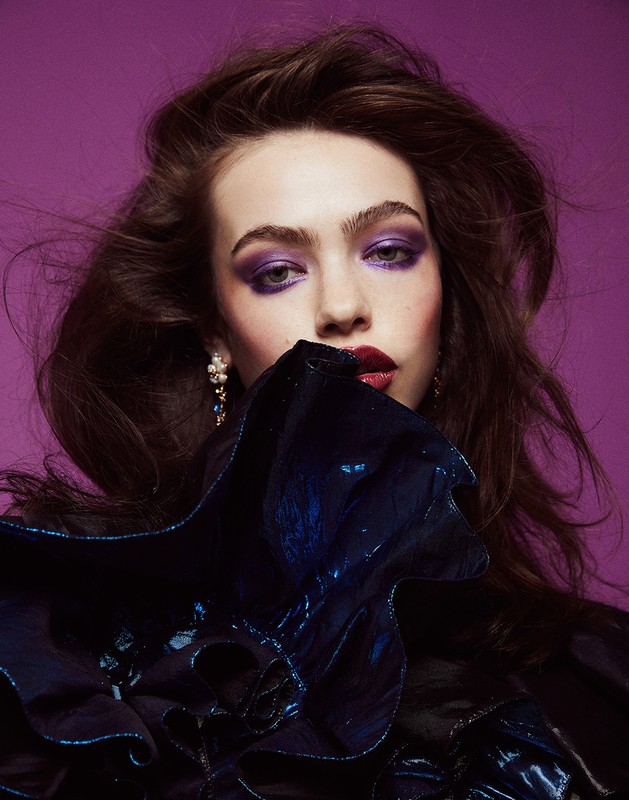 Close-up beauty portrait of a young woman with dark hair and dramatic purple makeup against a solid purple backdrop in a studio setting.