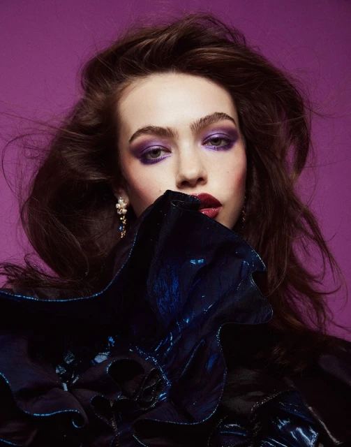 Close-up beauty portrait of a young woman with dark hair and dramatic purple makeup against a solid purple backdrop in a studio setting.