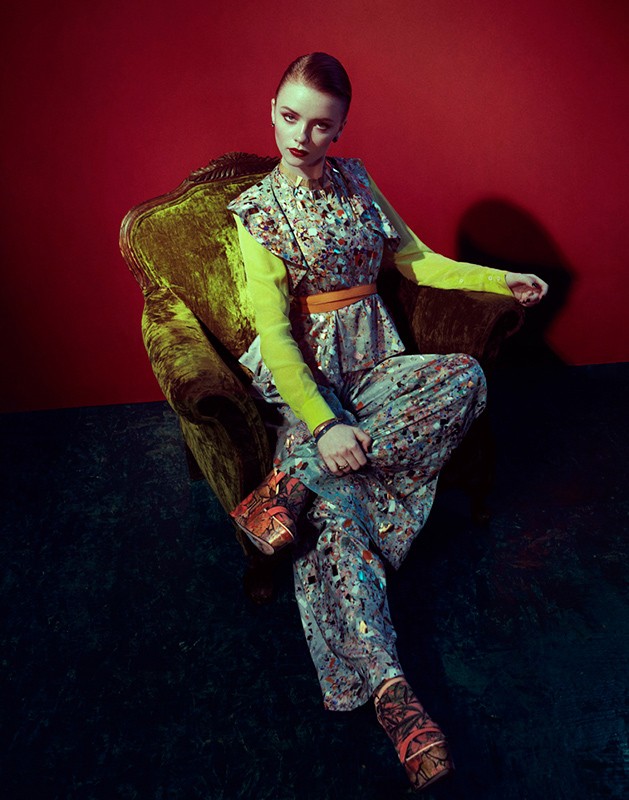 A young woman with red lipstick sits in a velvet chair wearing patterned pants and a top, with a bright yellow long-sleeve shirt underneath.