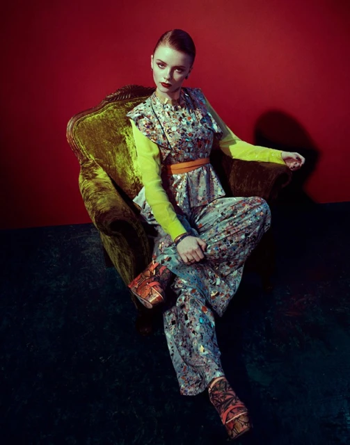 A young woman with red lipstick sits in a velvet chair wearing patterned pants and a top, with a bright yellow long-sleeve shirt underneath.