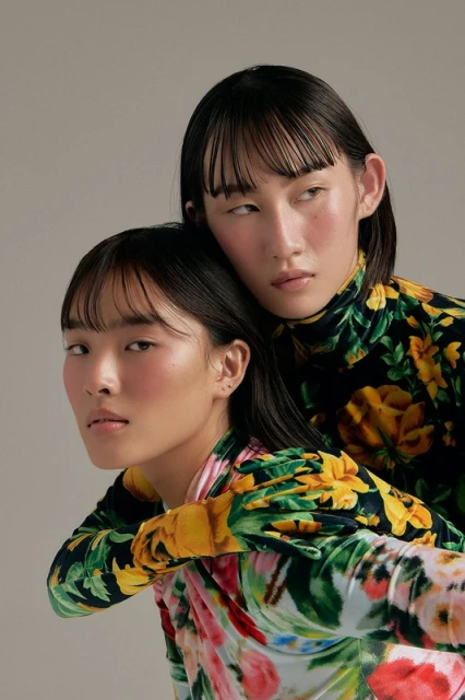 Two Asian women pose closely wearing floral print turtlenecks in a studio shot with a neutral background, fashion portrait.