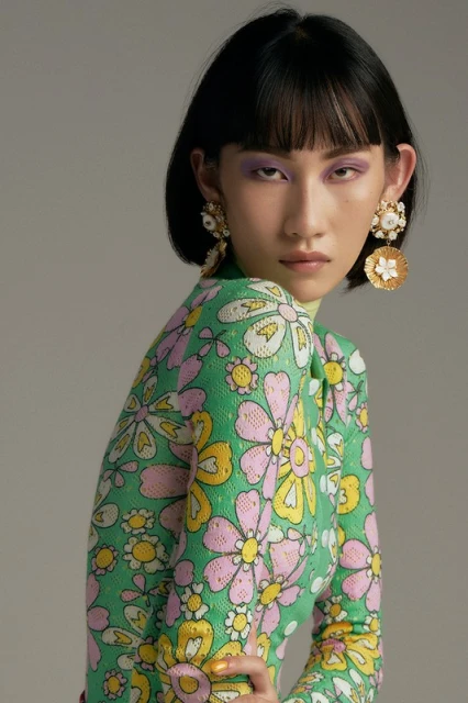 Woman with bob haircut wearing a floral print dress and statement earrings in a studio portrait with a neutral grey background.