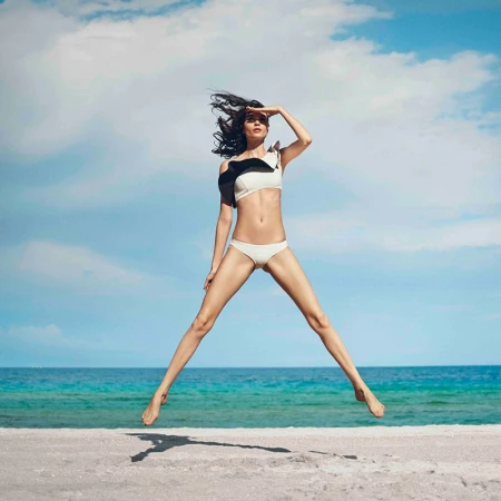 Woman in bikini jumps on beach with ocean and sky backdrop, hand shielding eyes, windblown hair, athletic figure, and summer vibes.