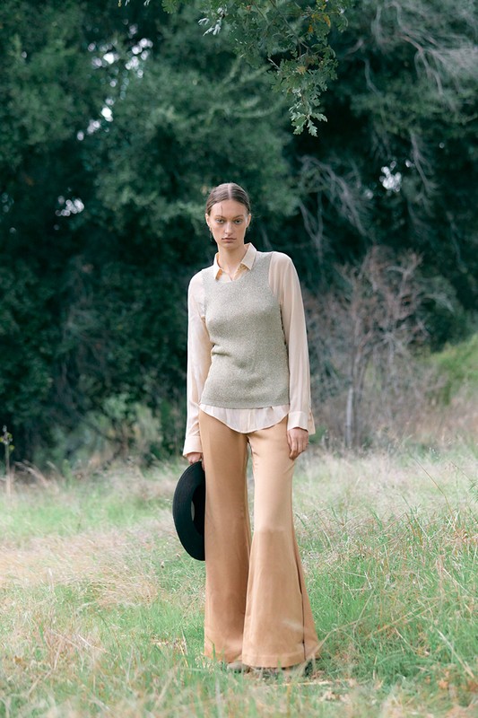 A stylish woman stands in a field wearing a top, vest, and bell-bottoms, holding a hat, surrounded by grass and trees.