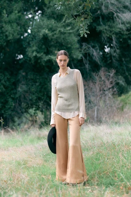 A stylish woman stands in a field wearing a top, vest, and bell-bottoms, holding a hat, surrounded by grass and trees.