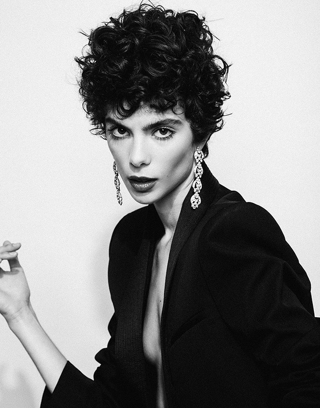 Close-up monochrome portrait of a woman with short dark curly hair wearing a dark blazer and dangling earrings