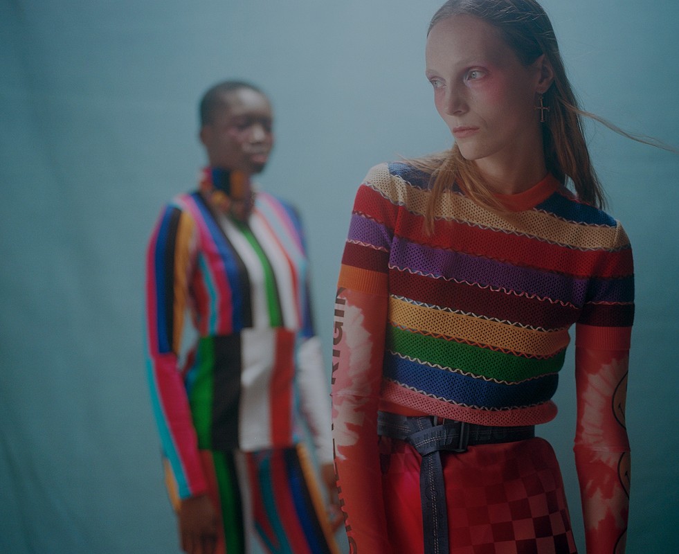 Two women in a studio setting, wearing colorful striped outfits, one with rainbow stripes and one with vertical color stripes.