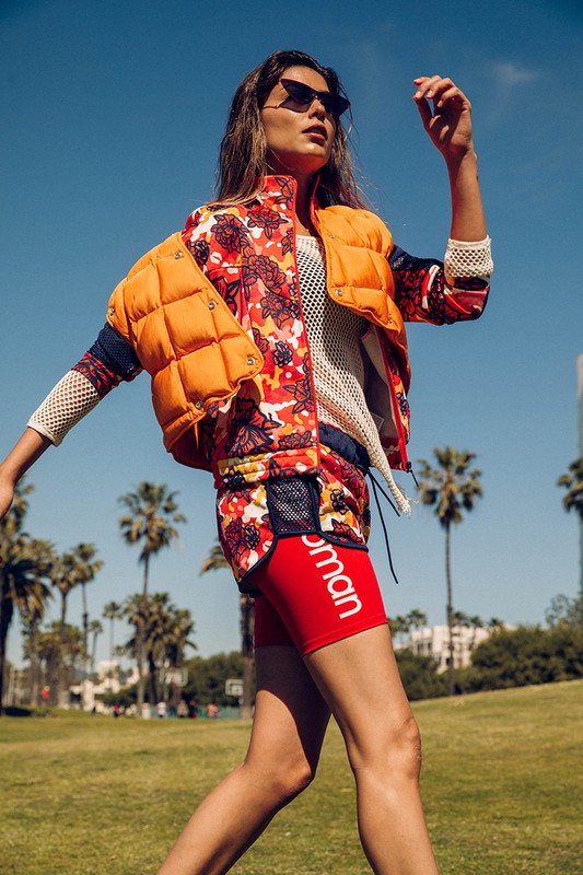 A woman walks on a grassy field in a floral print jacket and red athletic shorts, with palm trees and blue sky in the background.