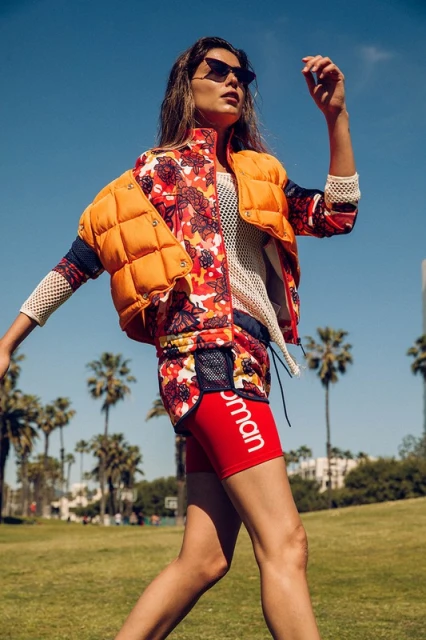 A woman walks on a grassy field in a floral print jacket and red athletic shorts, with palm trees and blue sky in the background.