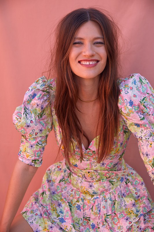 Portrait of a smiling woman with long brown hair wearing a floral dress against a light pink wall. Head and shoulders view, eye-level angle.