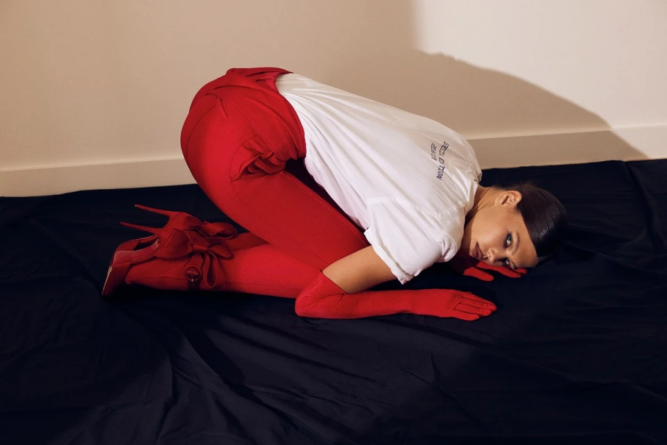 Brunette woman in a white t-shirt, red pants, gloves, and high heel boots, lying on a black surface in a brightly lit room.