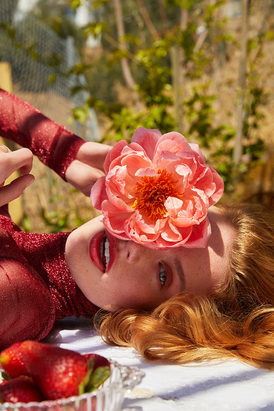 Red-haired woman in red lies on a white surface outside with strawberries, smiling with a pink flower covering one eye.
