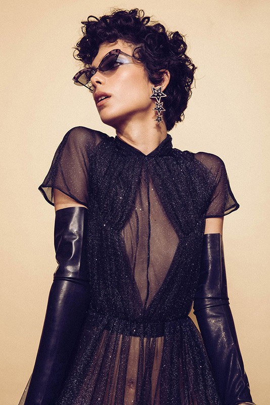 Fashion portrait of a woman with short curly hair wearing a black dress, long black gloves, statement star earrings, and sunglasses.