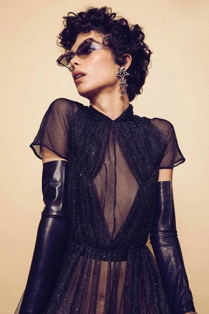 Fashion portrait of a woman with short curly hair wearing a black dress, long black gloves, statement star earrings, and sunglasses.