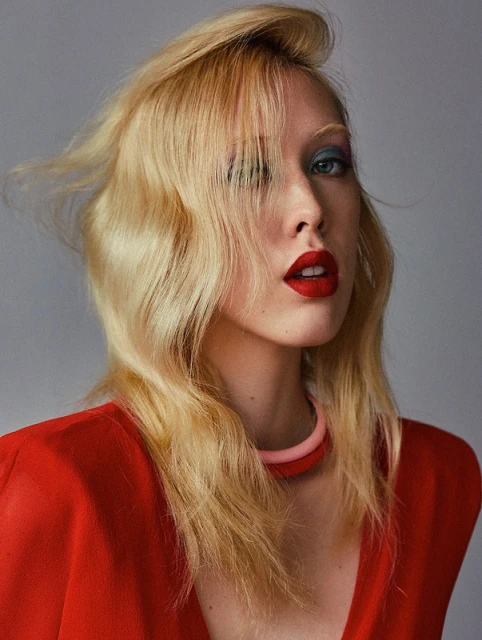 Portrait of a fair-skinned woman with blond hair wearing a red outfit and statement makeup with bright red lipstick against a grey background.