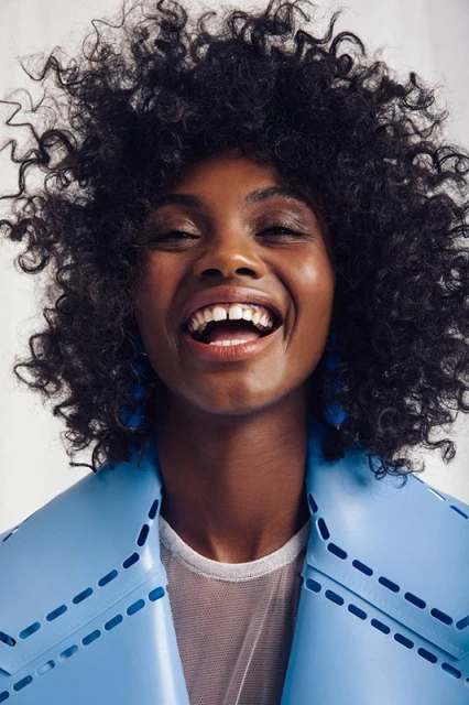 Portrait of a smiling Black woman with curly hair wearing a blue jacket and geometric earrings against a plain white backdrop.